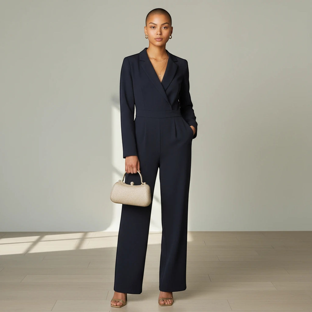 Modell bär en guldfärgad MiRA handväska med strukturerad form och metallhandtag till en elegant mörkblå jumpsuit.