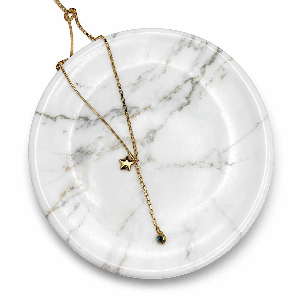 MiRA elegant guldfärgat Y-halsband med stjärnhänge och en liten gnistrande blå sten på ett vitt marmorfat.
