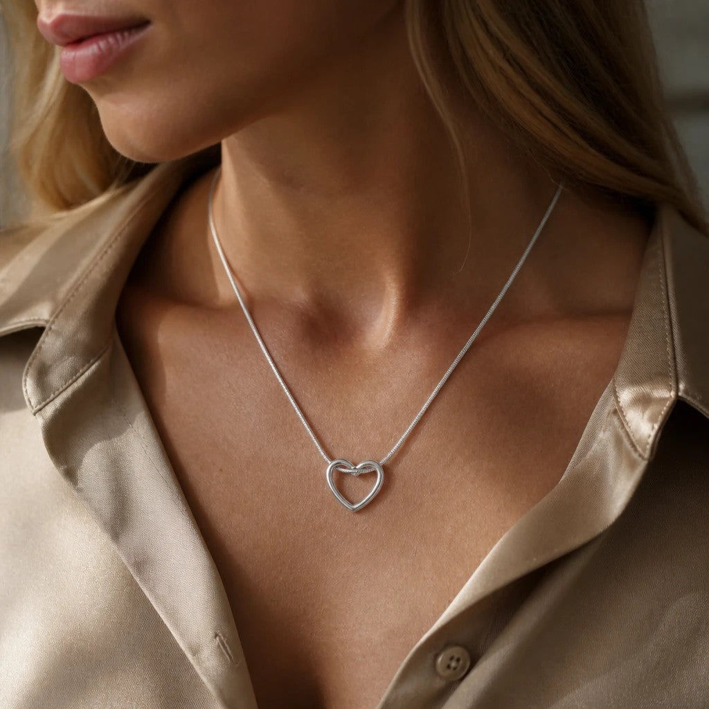MiRA halsband med öppet hjärtformat hänge i silverfärgad ormlänk på en kvinna i beige sidenblus.