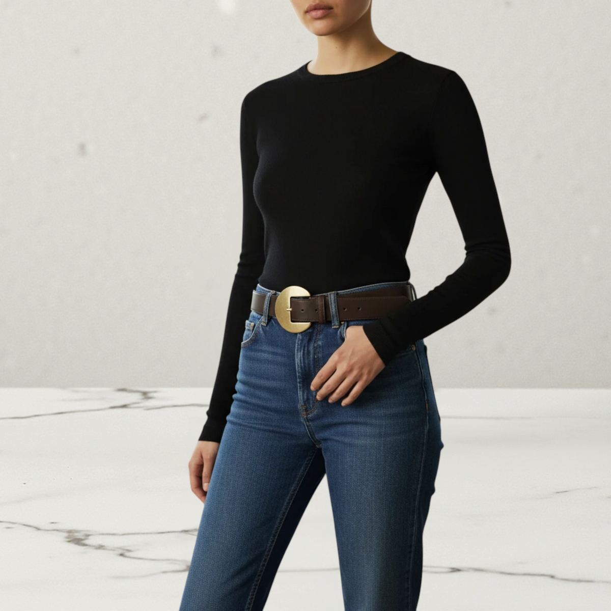 MiRA mörkbrunt damskärp i veganskt läder med ett runt guldfärgat spänne buret av en kvinna i jeans och svart tröja.