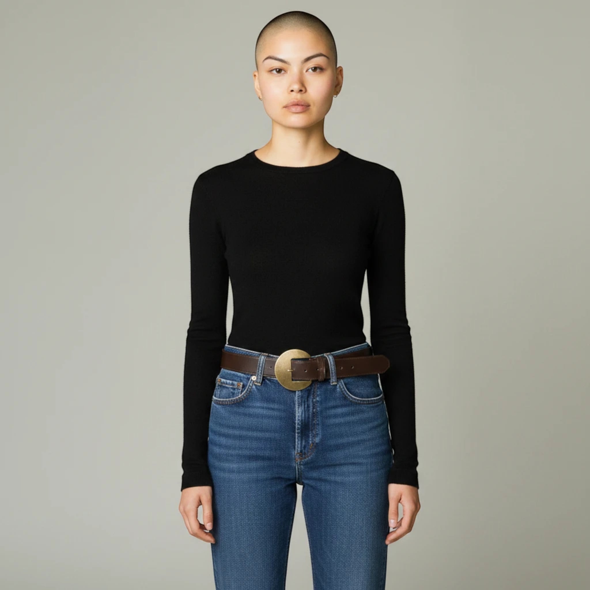 MiRA mörkbrunt damskärp i veganskt läder med ett runt guldspänne, buret av en modell i jeans och svart tröja.
